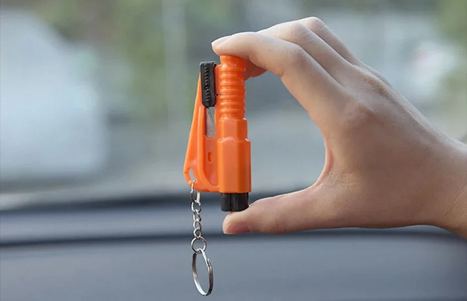 A hand holds an orange emergency car escape tool with a window breaker and seatbelt cutter attached to a chain.