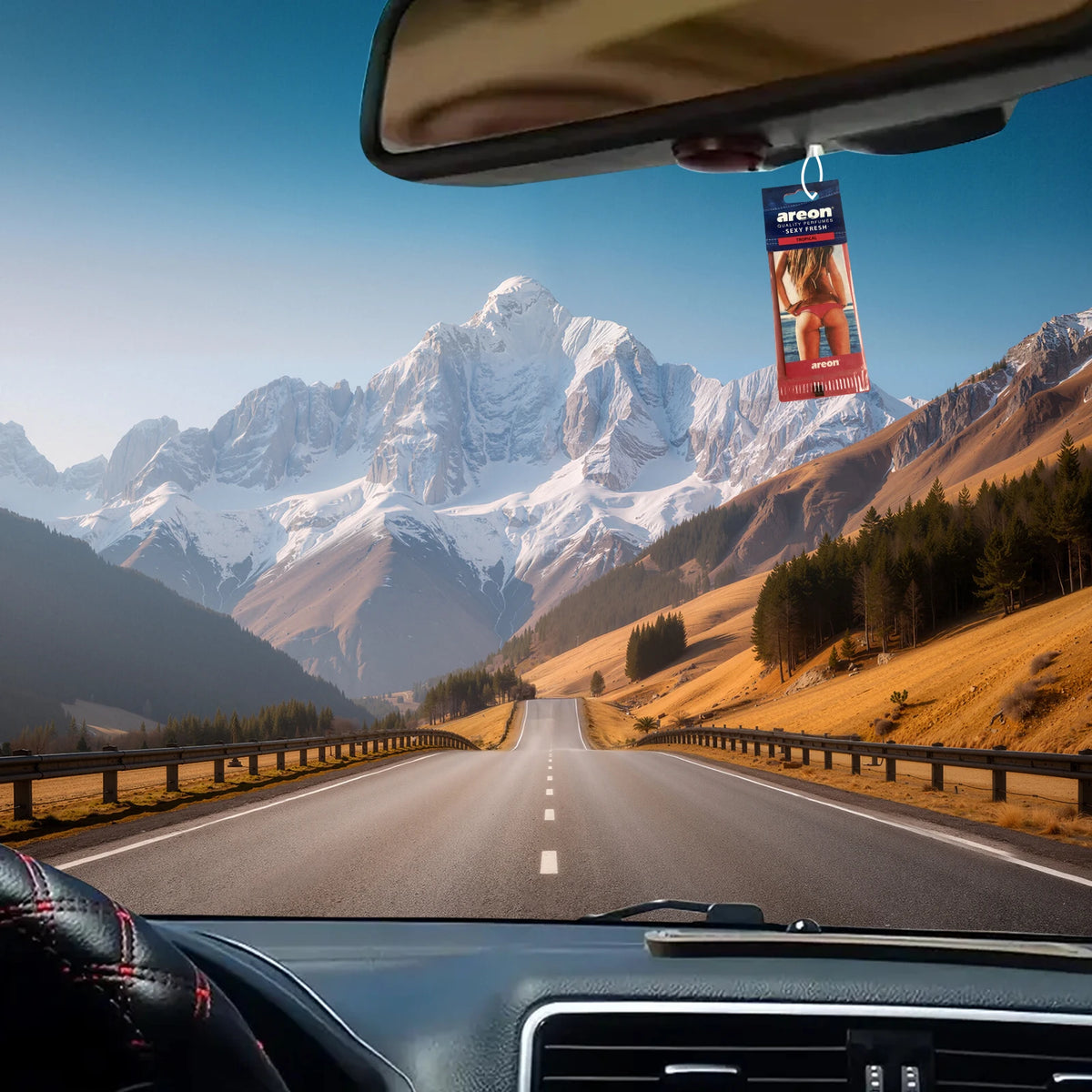 A multi-scent hanging air freshener is visible in the rearview mirror of a car driving on a scenic mountain road.