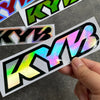 A hand holds a colorful racing suspension decal featuring the letters KYB on a dark gray surface.
