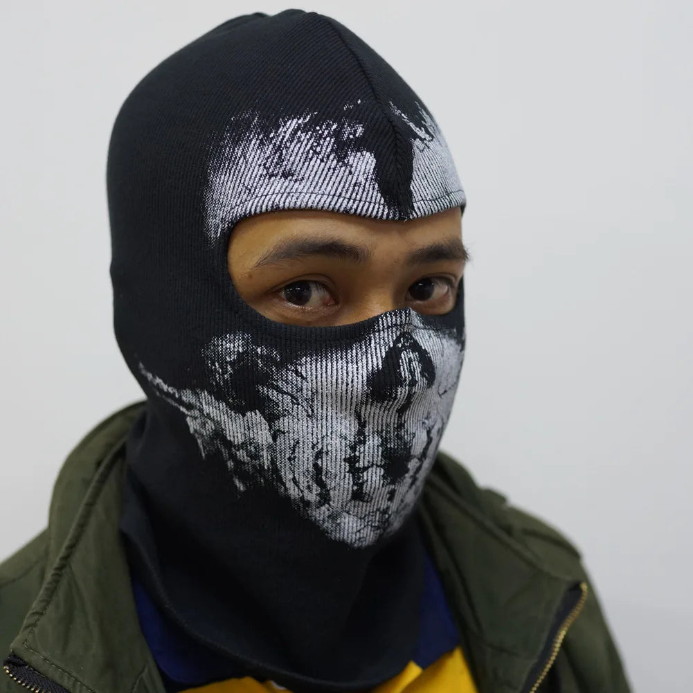 Una persona vistiendo una máscara balaclava negra con un patrón de cara desgastada blanca, visible bajo una chaqueta verde.