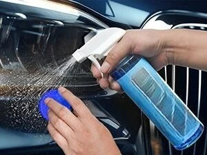 A hand is using a white spray nozzle to apply water onto a dark car surface for detailing.