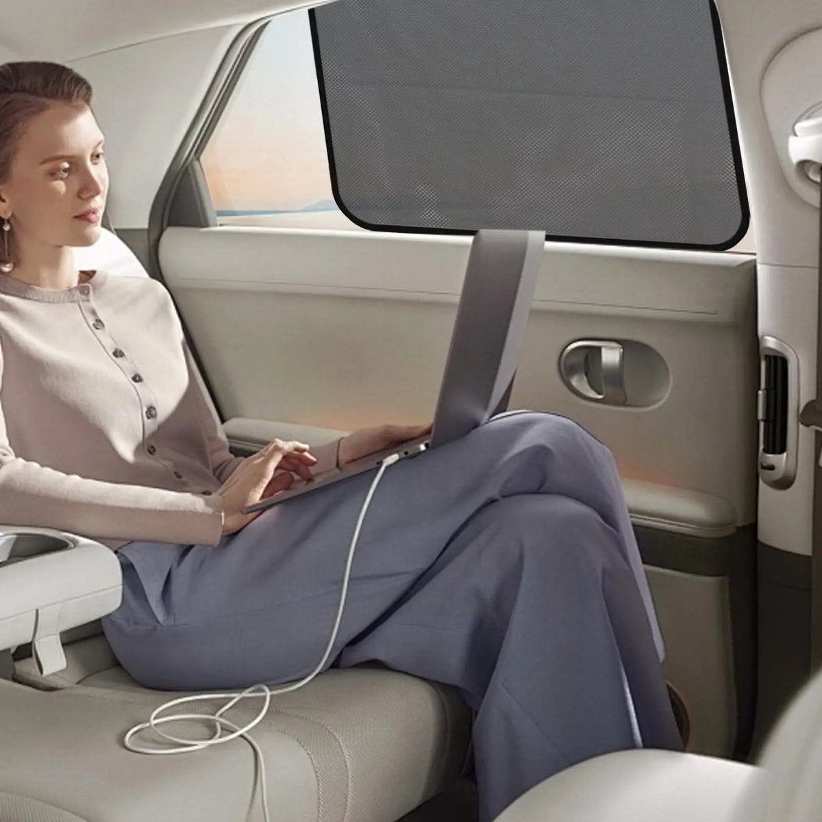 A woman sits in a car, using a laptop while a magnetic sunshade is visible on the window.