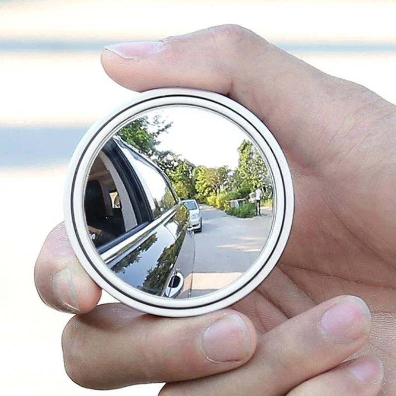 A hand holds a round convex blind spot rearview mirror reflecting an outdoor scene.