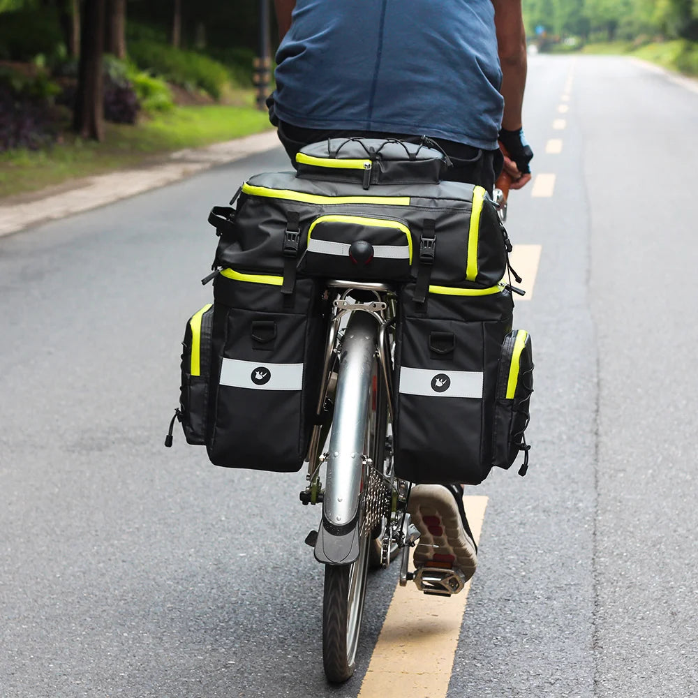 A person is riding a bicycle equipped with a black and neon yellow 3-in-1 mountain bike trunk bag set on an asphalt road.