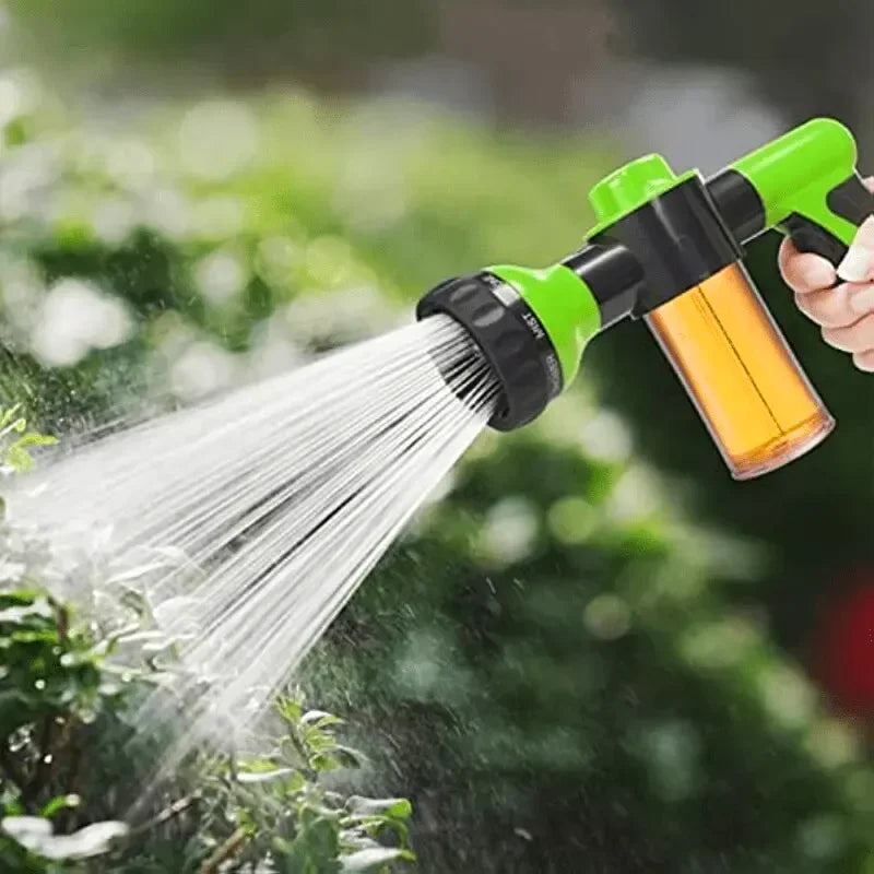 A hand is using a green and black high pressure foam lance car wash sprayer to spray water onto green foliage.