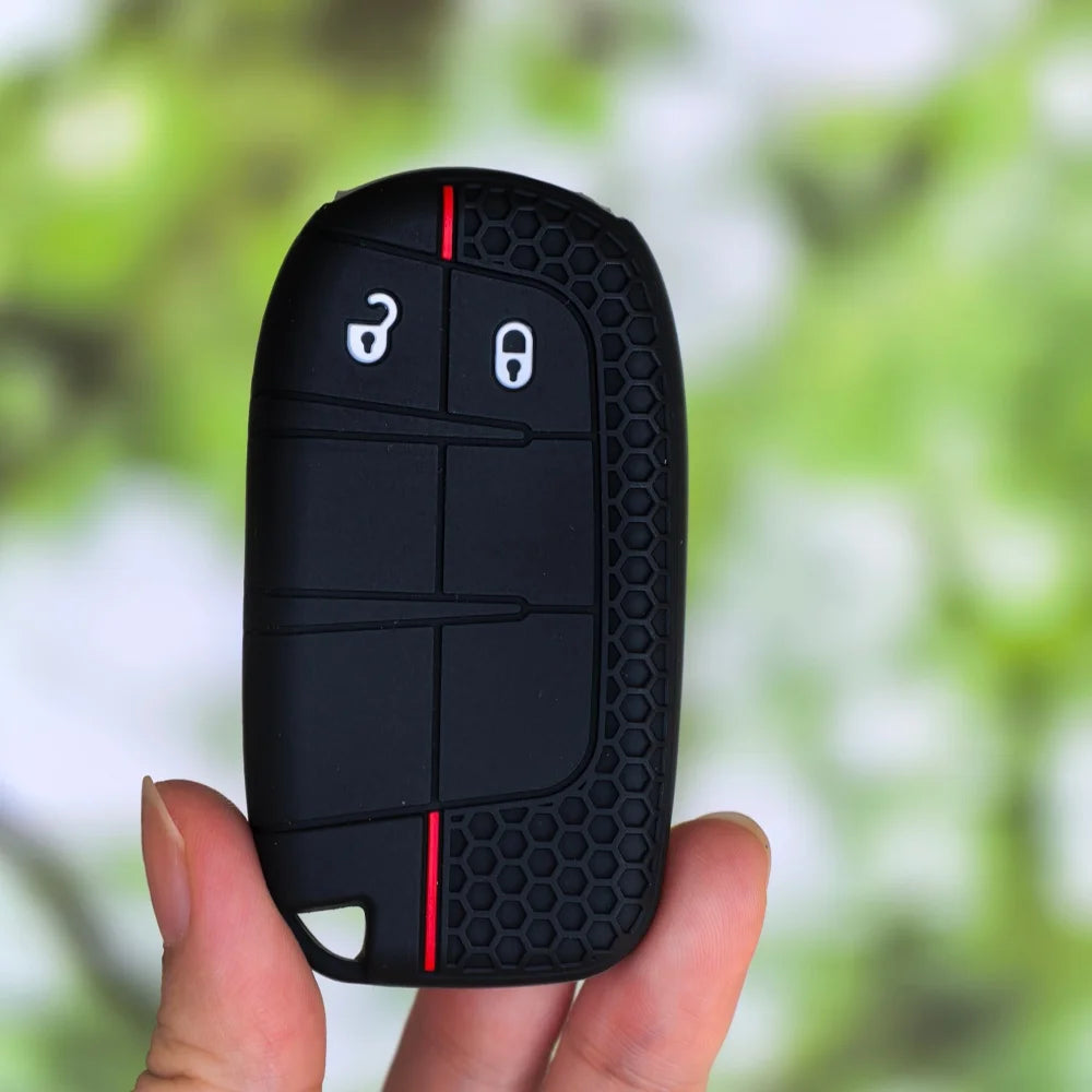 A black silicone car key cover for Jeep, Dodge, and Chrysler models is held in a hand against a blurred green background.