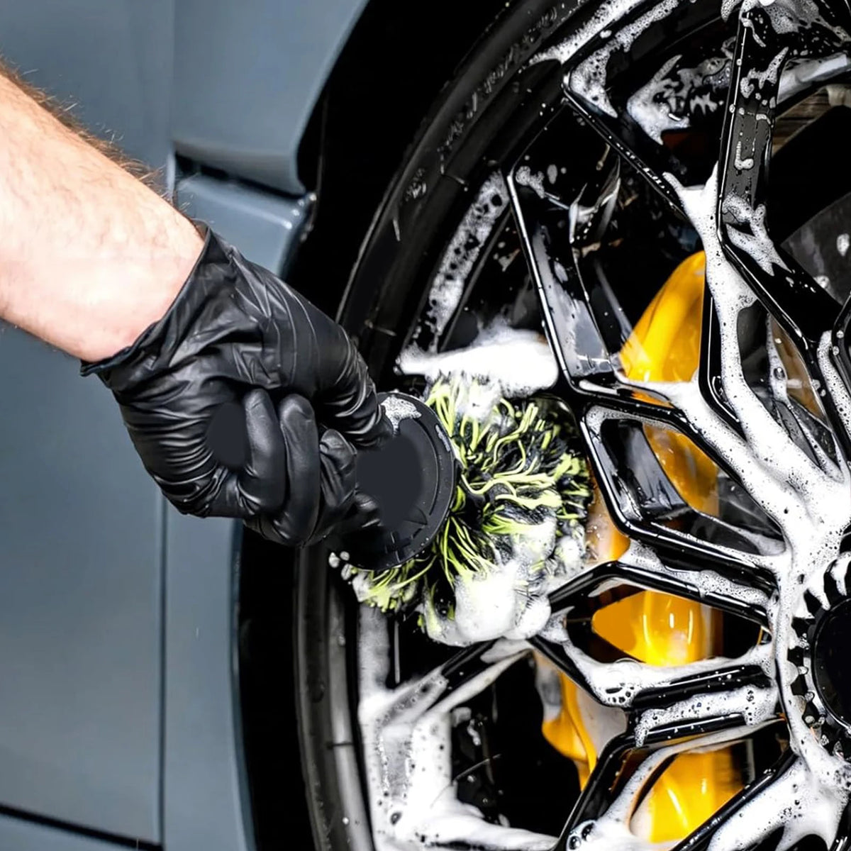 A hand wearing a black glove is using a heavy-duty wheel barrel brush to clean the spokes of a black car wheel.