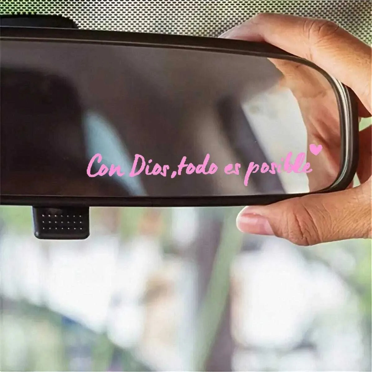 A hand holds a black car mirror with a pink heart decal that reads Con Dios todo es posible
