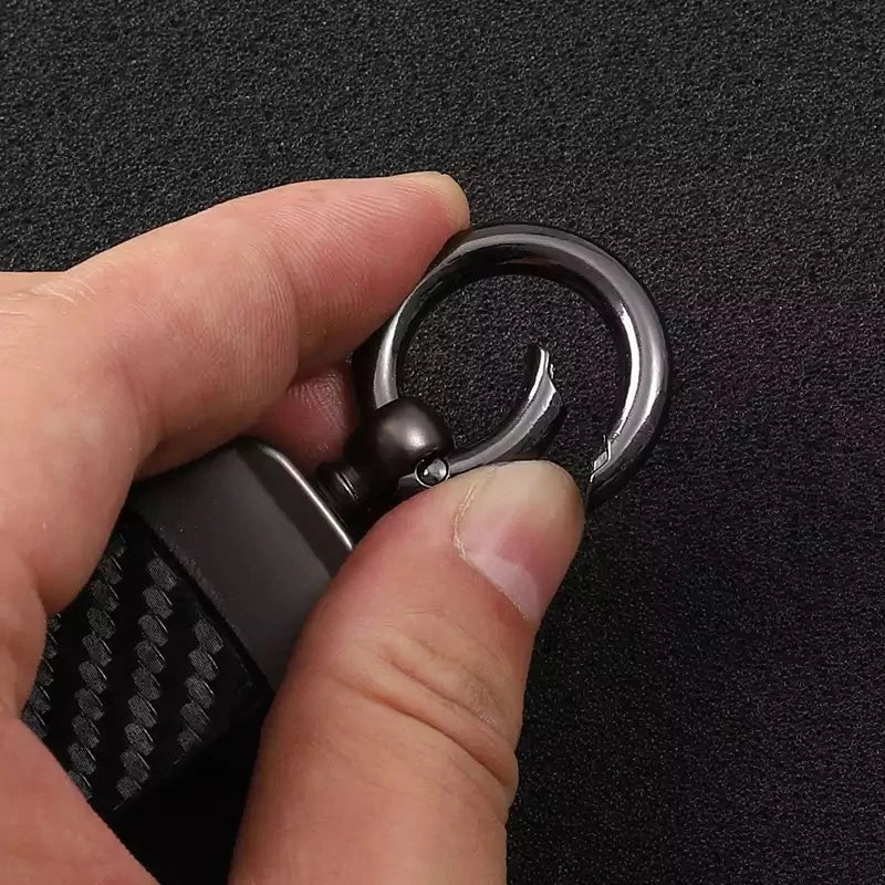 A hand holds a black and silver keychain with a carbon fiber texture and a metal ring.