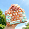 A hand holds a colorful keycap stress relief button set featuring cute cartoon designs against a bright blue sky.