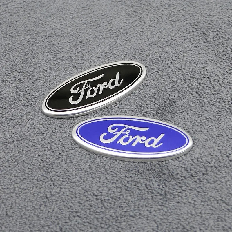 Zwei ovale Ford-Embleme, eines schwarz und eines blau, liegen auf einer grauen, strukturierten Oberfläche.