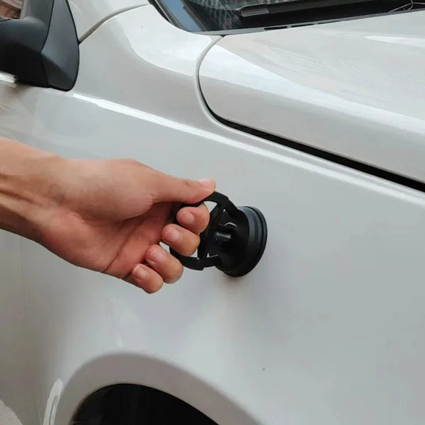 A hand is operating a black dent puller tool on the white body panel of a car.