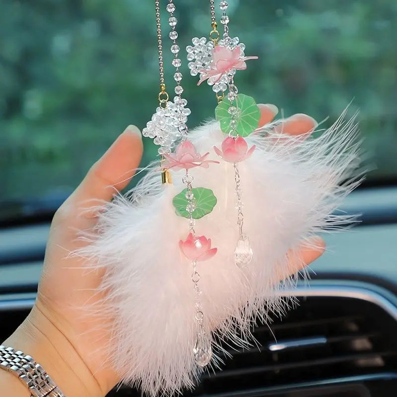 A hand holds a pink lotus car pendant ornament featuring clear crystals and green leaves on a white feather background.