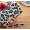 A hand holds a black and white leopard print long wallet on a wooden table next to some flowers.