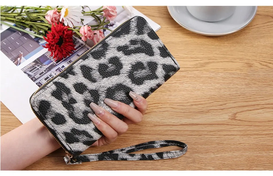A hand holds a black and white leopard print long wallet on a wooden table next to some flowers.