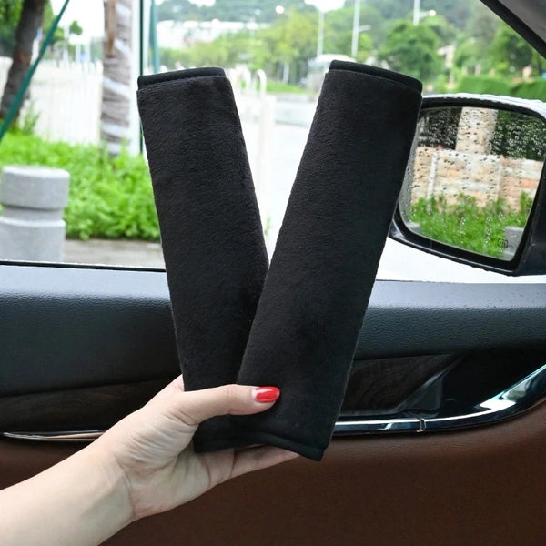 A hand holds two black plush seat belt covers for car and shoulder pads.
