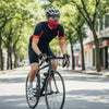 A man wearing a black cycling outfit and a red face mask rides a bicycle down a tree-lined street.