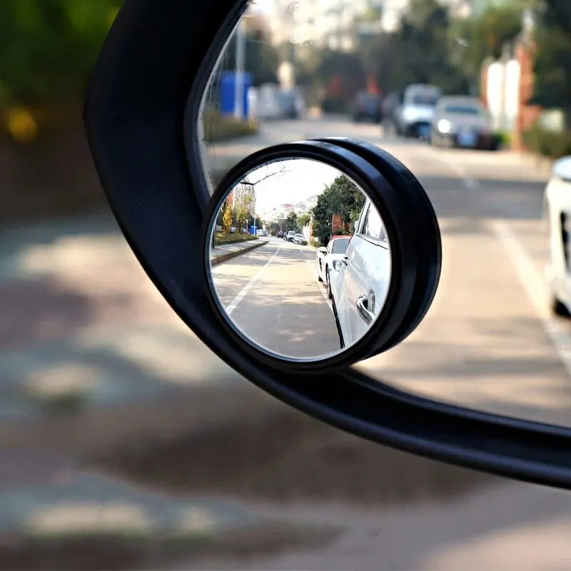A close-up view of a black car blind spot mirror set reflecting a street and a white car.