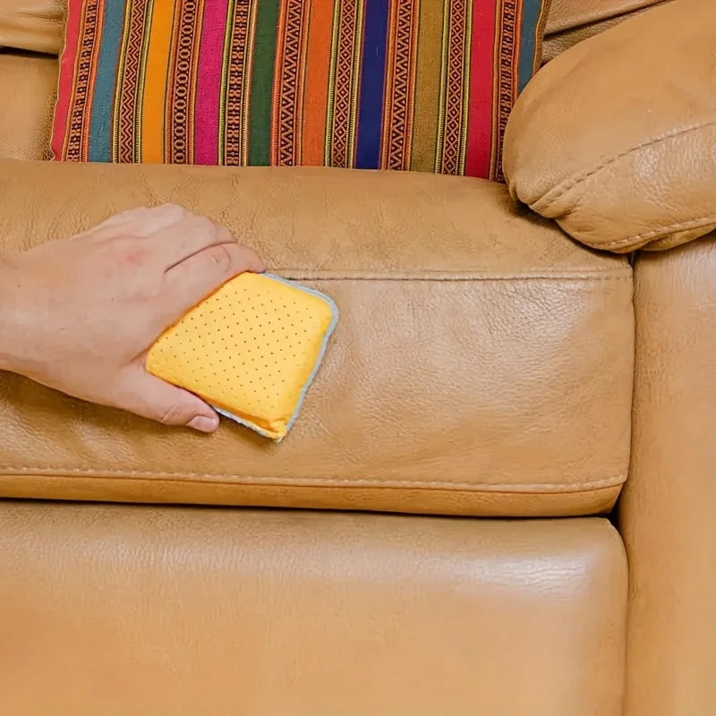 A hand is holding a yellow anti-fog glass and mirror cleaning sponge on a tan leather sofa.