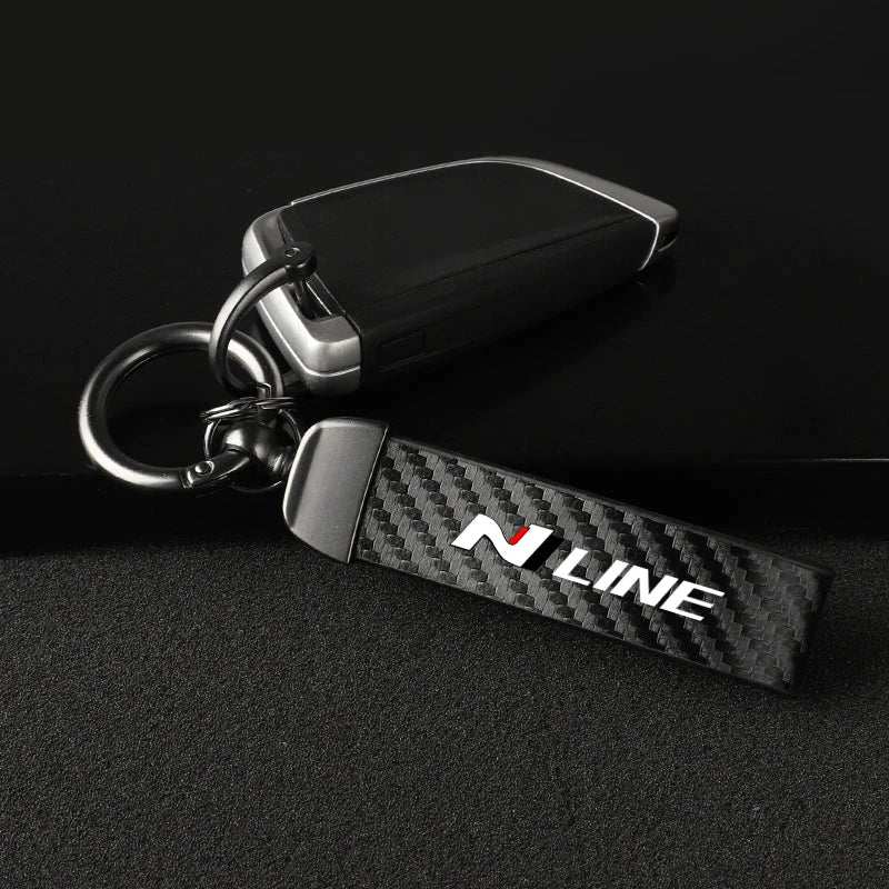 A black and carbon fiber style keychain with a silver metal fob and a black leather strap.