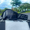 A pair of black off-road motorcycle goggles resting on a gray surface with greenery in the background.