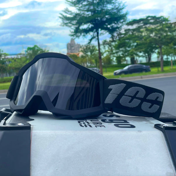 A pair of black off-road motorcycle goggles resting on a gray surface with greenery in the background.