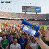 A hand holds a blue portable air horn, with a crowd visible in the background.