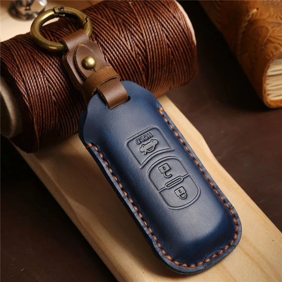 A dark blue leather car key fob cover attached to a brass keyring, resting on a wooden surface next to a rolled brown leather