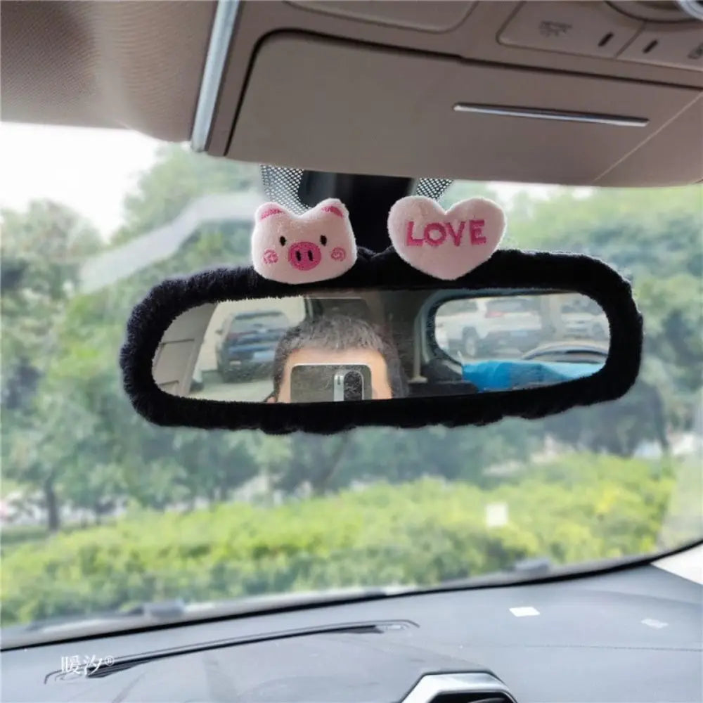 A rearview mirror cover decorated with pink pig and heart charms on a black fuzzy background.