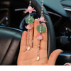 A hand holds several pink and green lotus-shaped car pendants decorated with clear crystals.