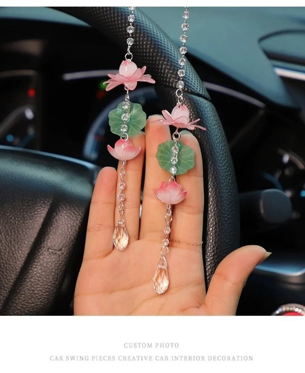 A hand holds several pink and green lotus-shaped car pendants decorated with clear crystals.