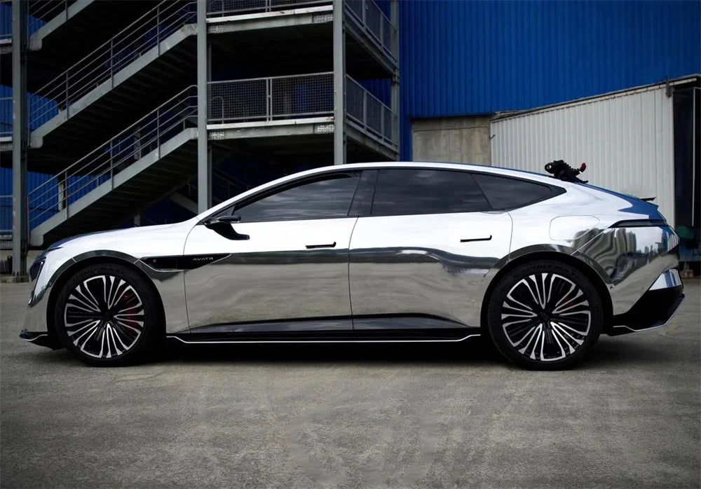 A silver car with a glossy chrome vinyl wrap is parked outdoors in front of an industrial building.