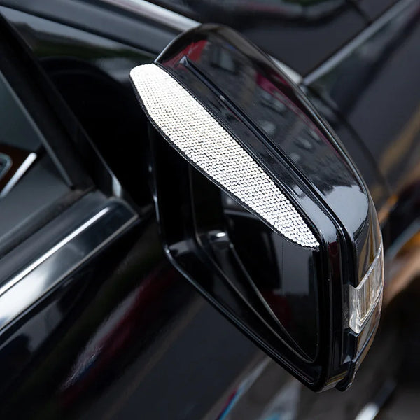 A close-up of a black car's rearview mirror with a rhinestone rain shield visor attached.