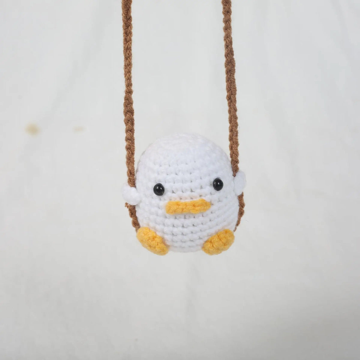 A white crocheted duck hanging from a brown string on a white background.
