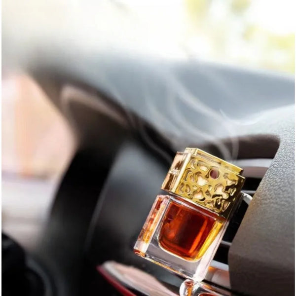 A glass bottle of car air freshener with a gold cap is placed in the vent of an air conditioning system.