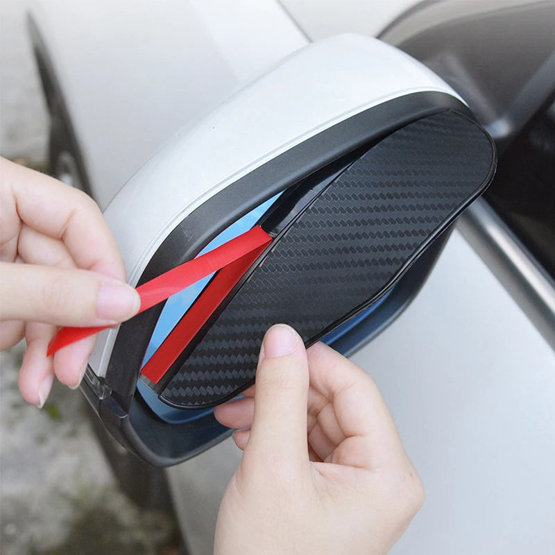 A hand is inserting a red and blue rain shield into the carbon fiber car rearview mirror.