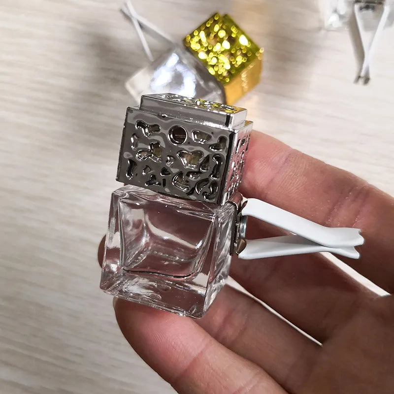 A clear glass car air freshener bottle with a silver mesh top and a white dispenser is held in a hand.