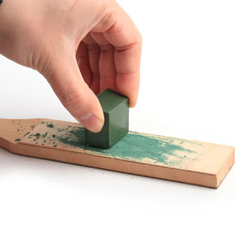 A hand presses a green metal polishing wax bar into a wooden block covered with green paint.