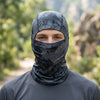 A man wearing a dark camouflage stretchy balaclava cap in an outdoor setting.