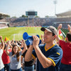 A young man holds a blue portable air horn, cheering in a crowd at an outdoor sporting event.