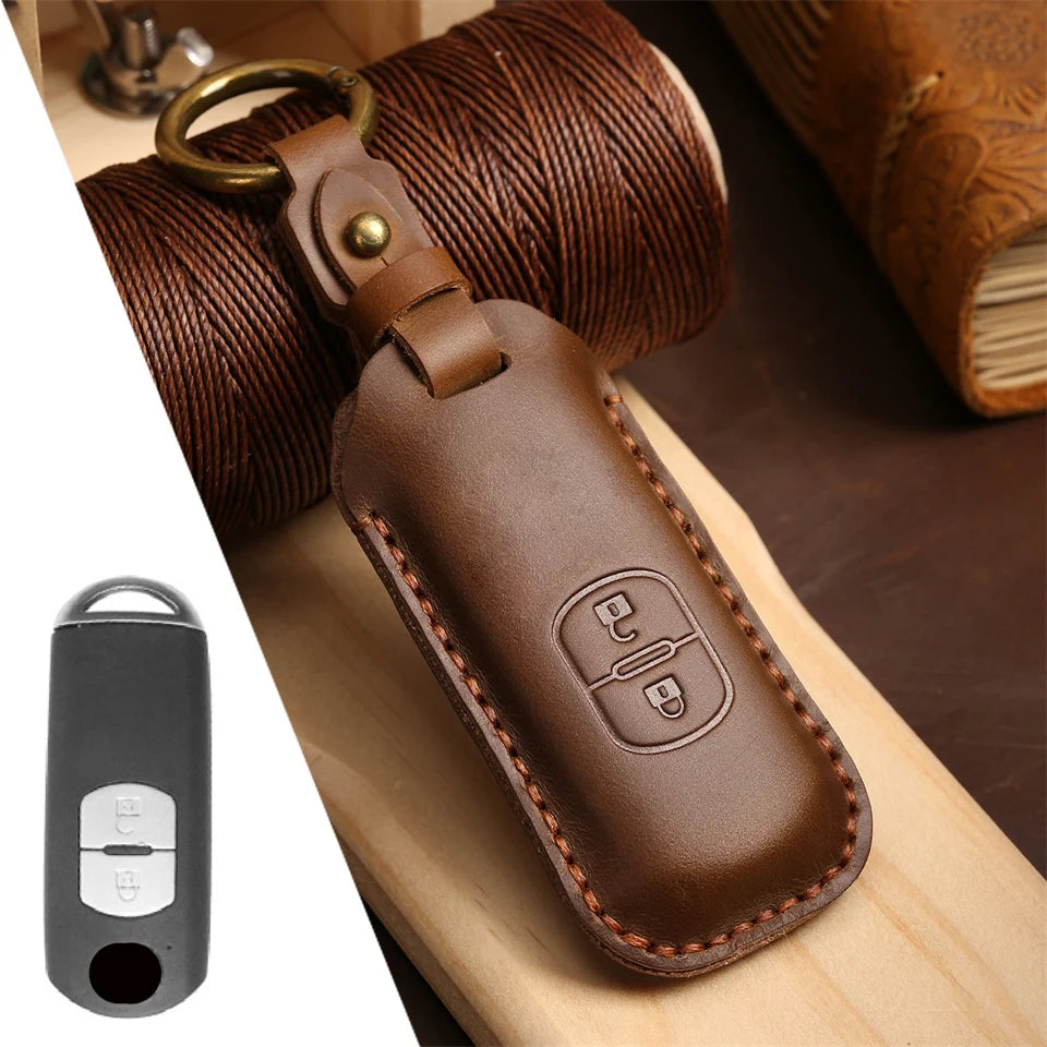 A brown leather car key fob cover is shown resting on a wooden surface next to a rolled-up brown leather strap.