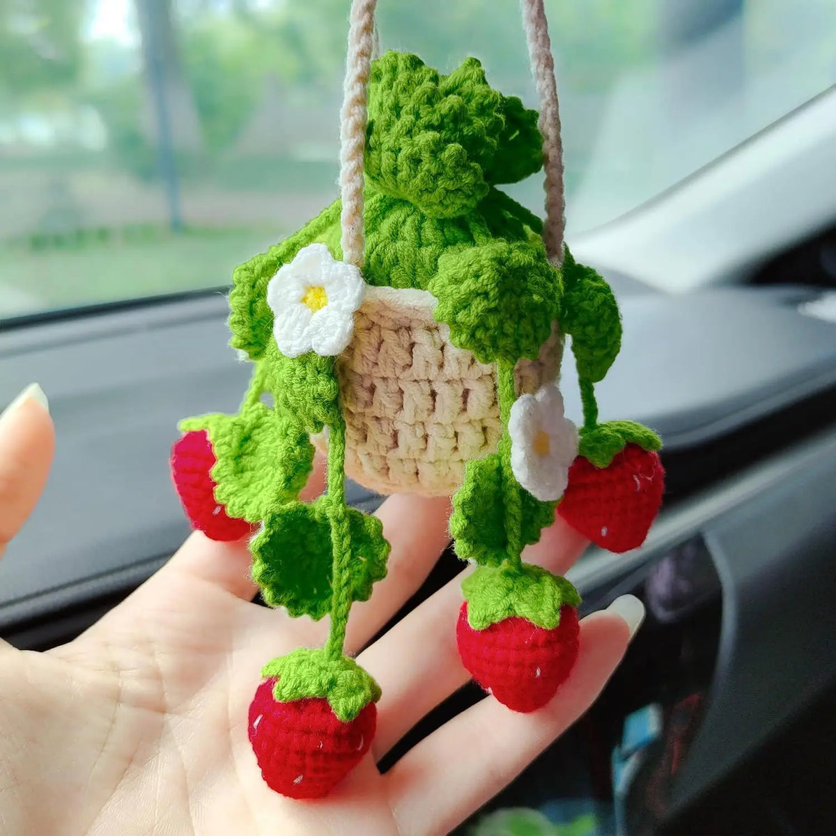 A handmade strawberry crochet hanging decor featuring green leaves and white flowers, displayed in a hand.