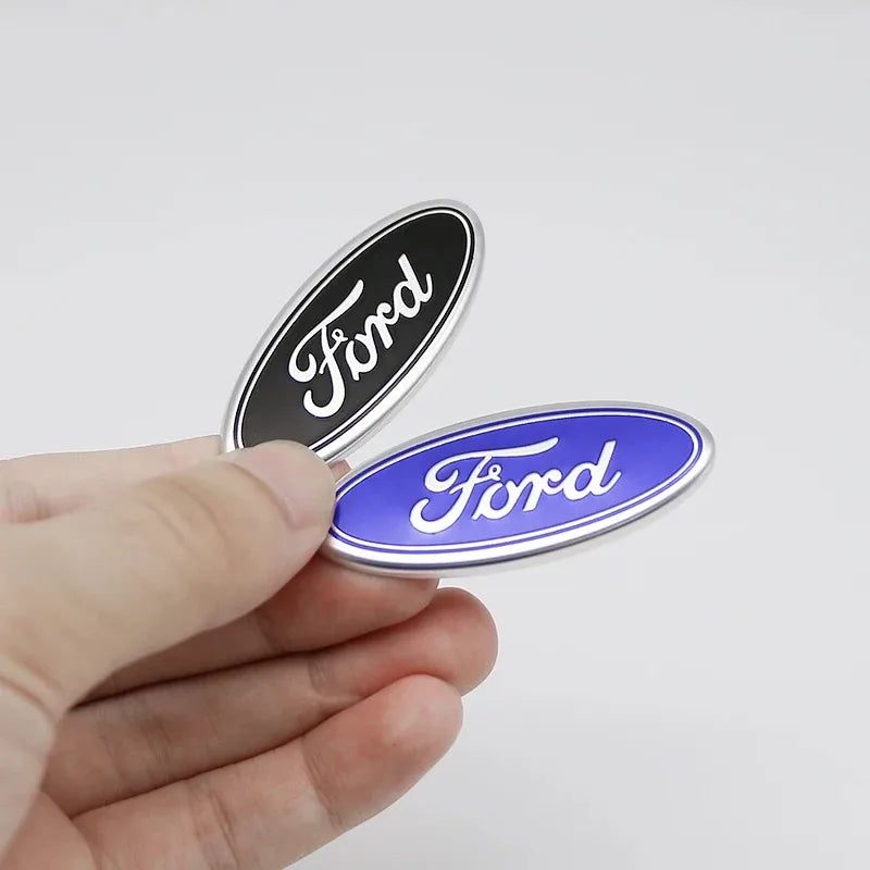 Eine Hand hält zwei ovale Ford-Lenkrad-Zieraufkleber, einer schwarz und einer blau.