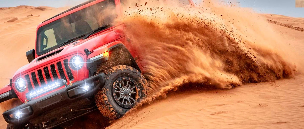 A red Jeep Wrangler driving through sand, kicking up a large cloud of orange dust.