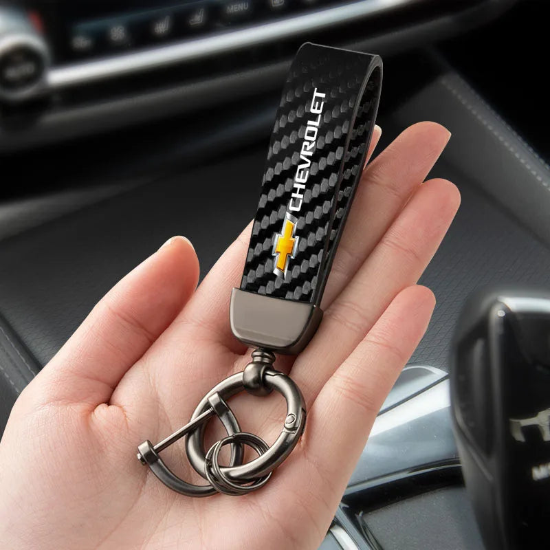 A hand holds a luxury metal keyring set with a black carbon fiber design and a gold Chevrolet emblem.