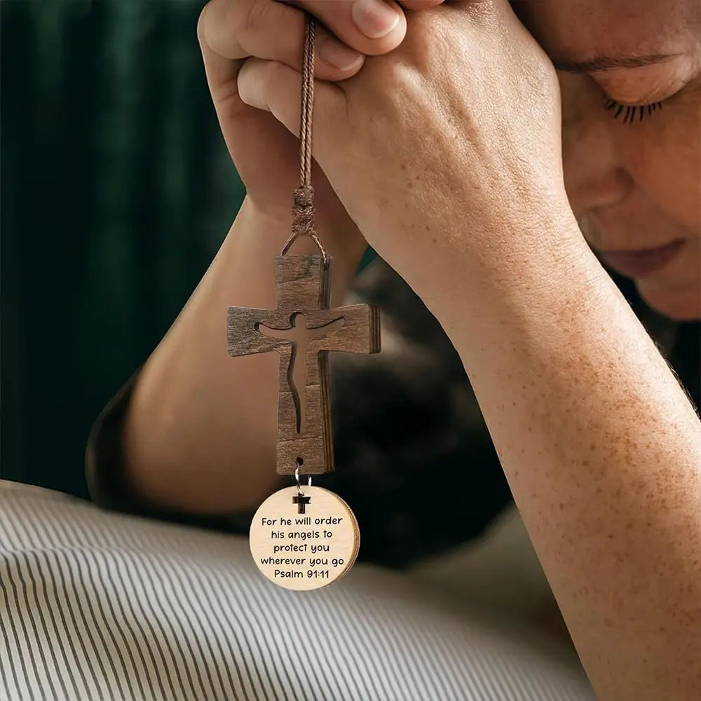 A hand holds a wooden cross ornament with an engraving on it, set against a blurred green background.