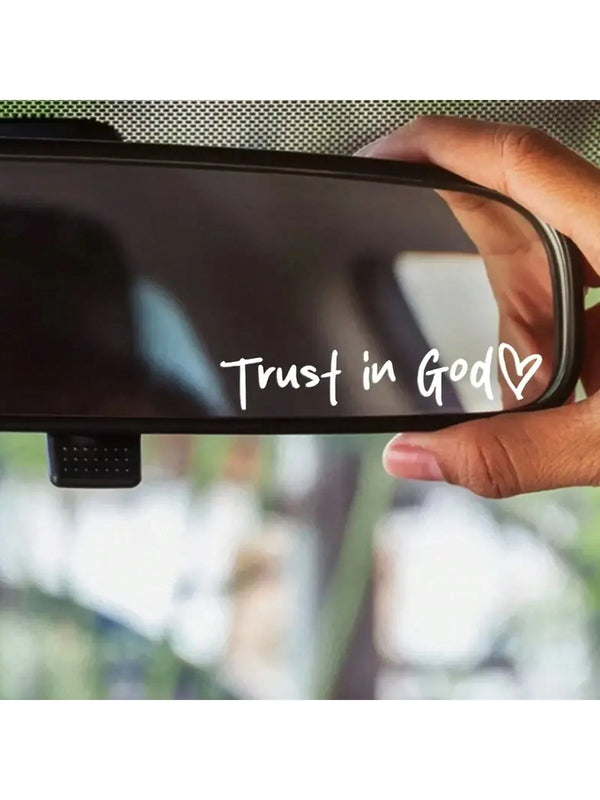 A hand holds a black car decal with the text Trust in God and a heart symbol on an interior mirror.