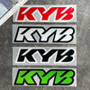 Four rectangular decals featuring the KYB logo in red, black, white, and neon green on a gray surface.