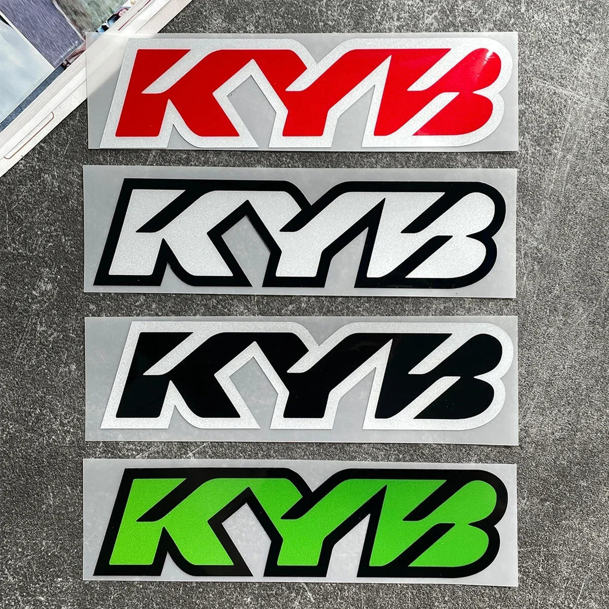 Four rectangular decals featuring the KYB logo in red, black, white, and neon green on a gray surface.