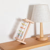 A wooden rainmaker musical toy with a clear tube and colorful beads sits next to a light-colored lampshade.